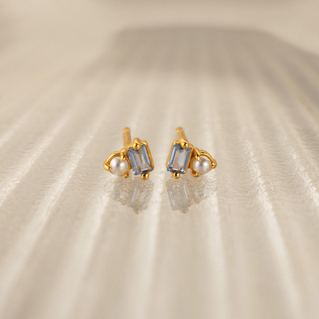 Pearl Aquamarine Studs feature rectangular aquamarine gemstones and small pearls set in gold, elegantly displayed on a textured beige surface—perfect for any occasion.