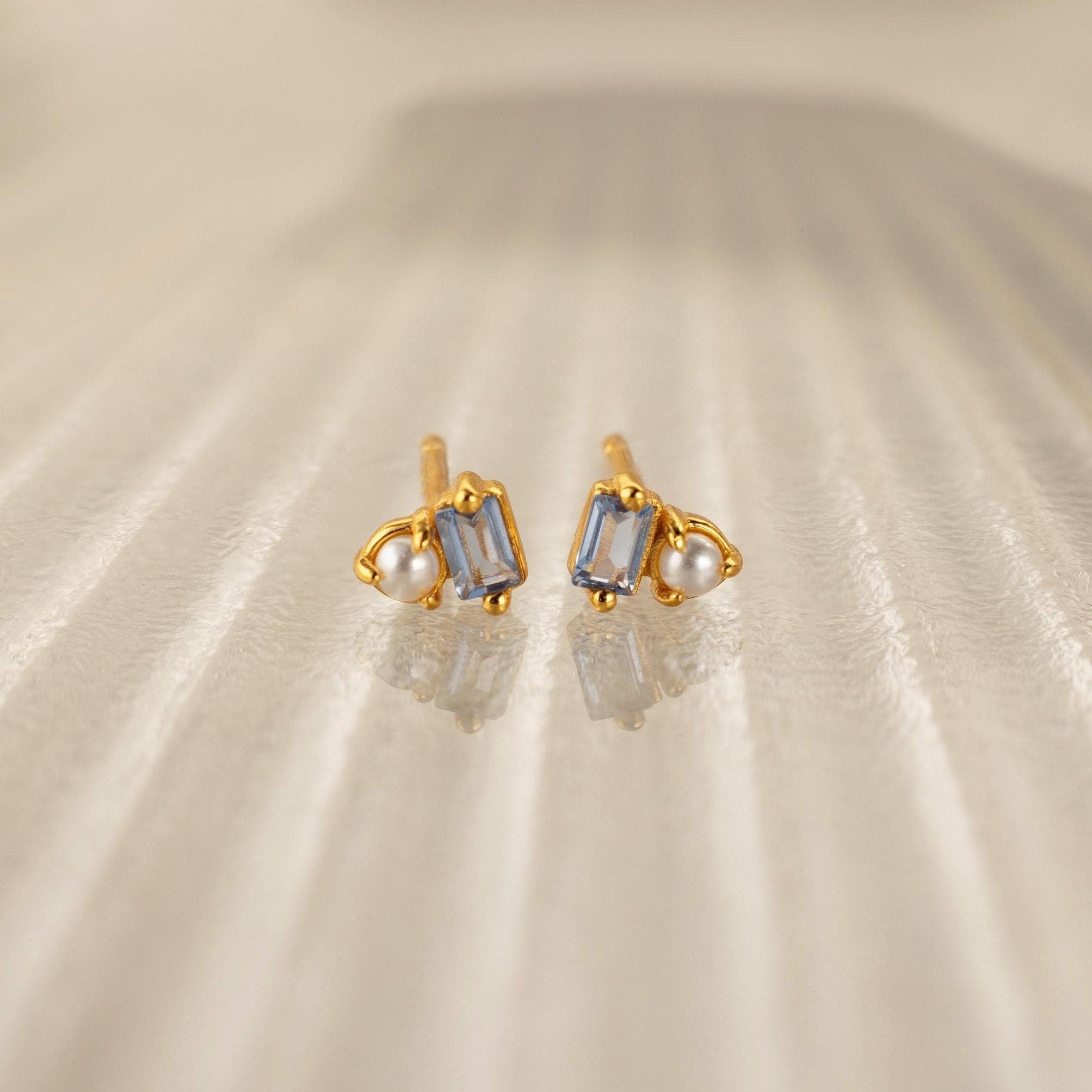 Pearl Aquamarine Studs feature rectangular aquamarine gemstones and small pearls set in gold, elegantly displayed on a textured beige surface—perfect for any occasion.