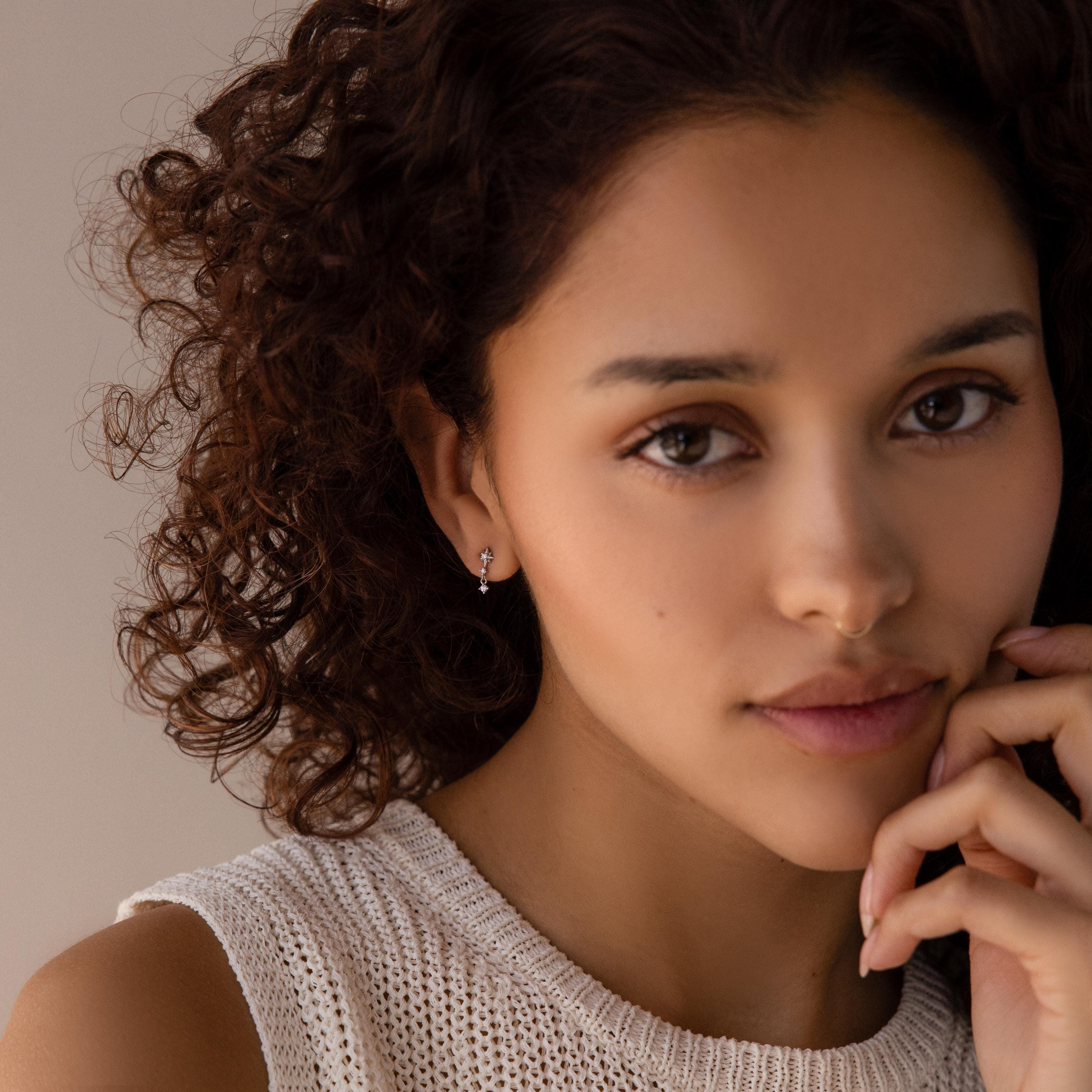 A woman with curly hair in a sleeveless knit top softly touches her face and gazes at the camera, her style enhanced by Diamond Starburst Drop Studs.