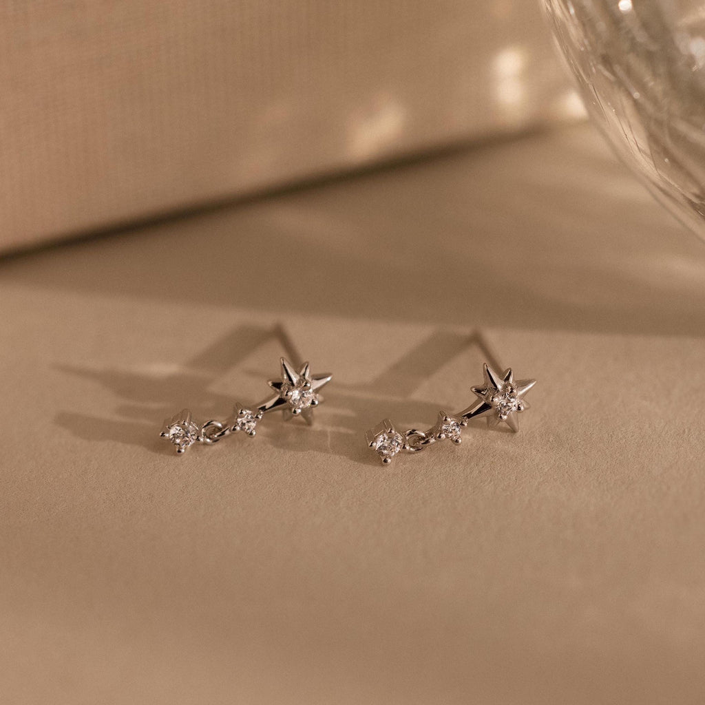 Diamond Starburst Drop Studs in Sterling Silver, star-shaped earrings with sparkling gems, are displayed on a neutral beige surface.