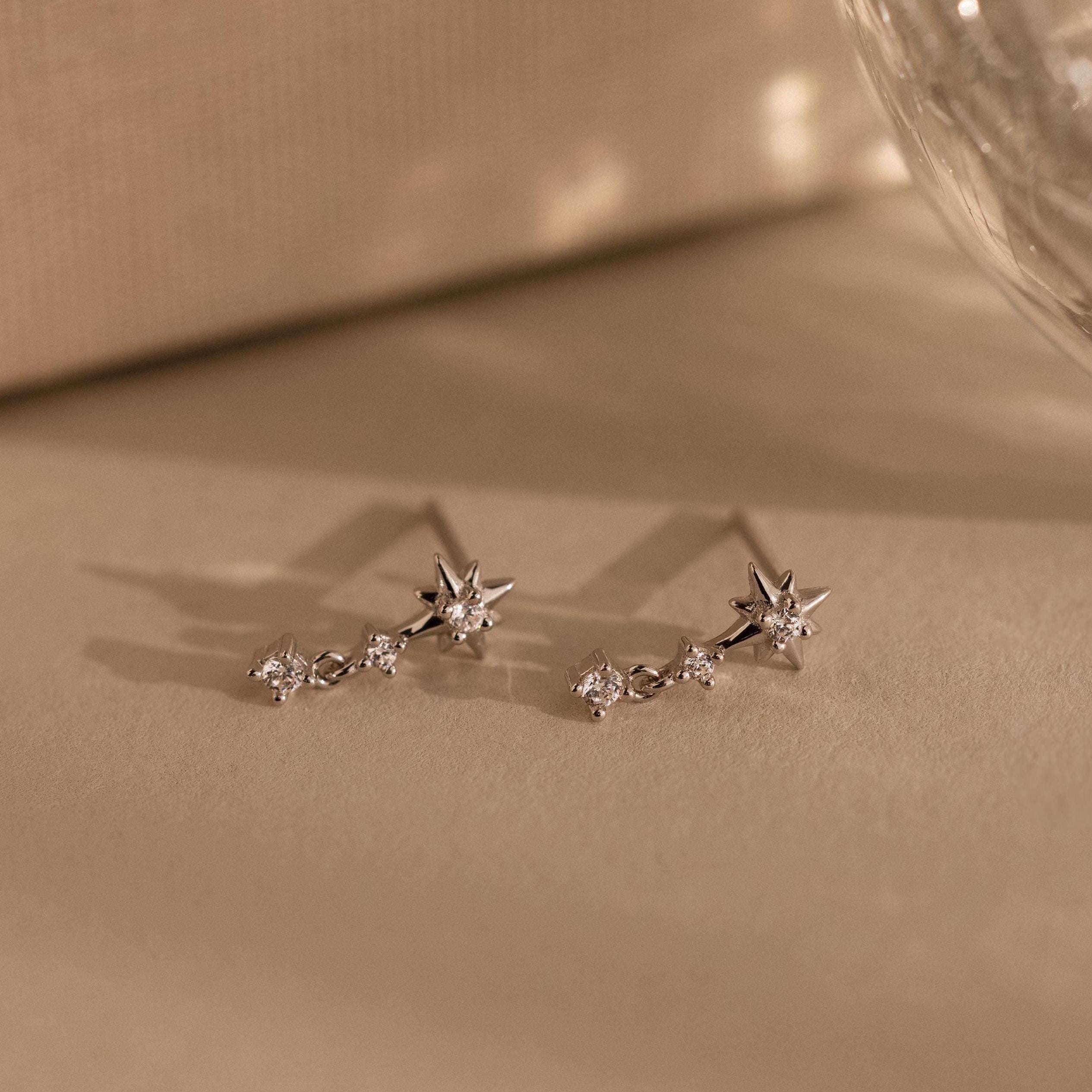 Diamond Starburst Drop Studs in Sterling Silver, star-shaped earrings with sparkling gems, are displayed on a neutral beige surface.