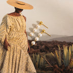 A woman in a yellow patterned dress and straw hat elegantly showcases the Dainty Pearl Drop Studs.