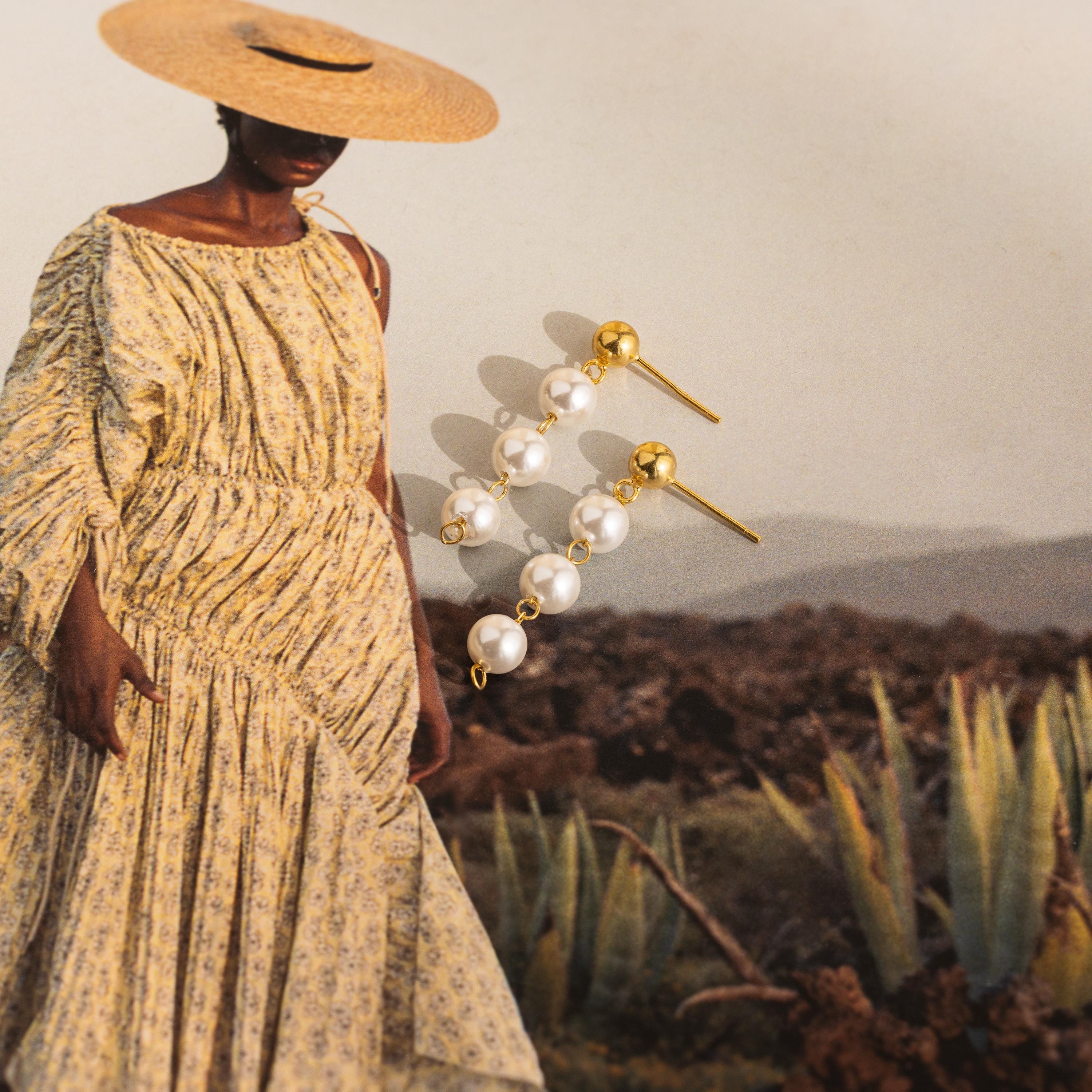 A woman in a yellow patterned dress and straw hat elegantly showcases the Dainty Pearl Drop Studs.
