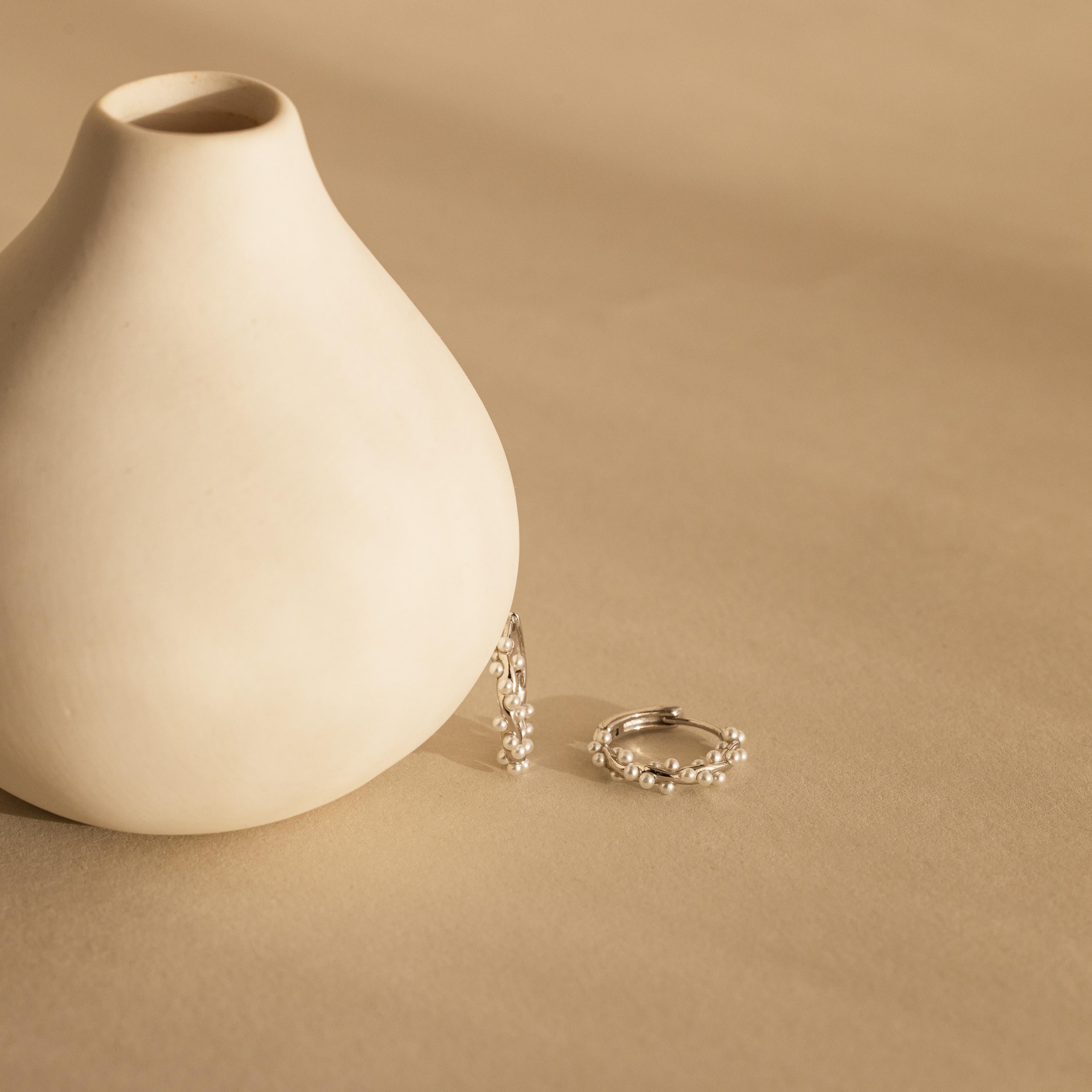 The Pearl Cluster Huggies in Sterling Silver, featuring small pearls, rest beside a smooth white ceramic vase on a beige surface—perfect as elegant bridal earrings or for any special occasion.