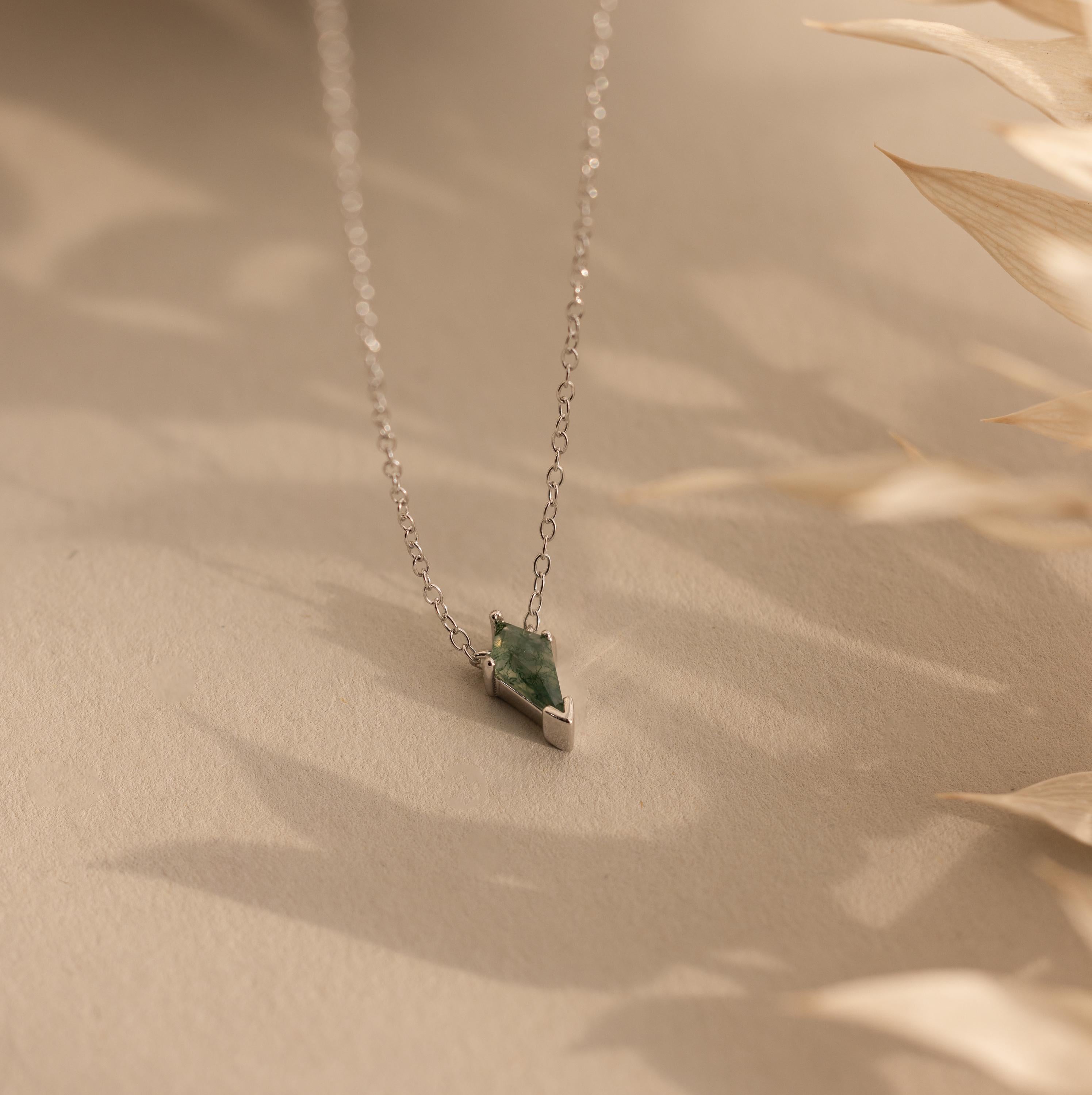 The Agate Art Deco Pendant Necklace, crafted in delicate silver and featuring a moss agate pendant, is elegantly displayed on a beige surface with soft shadows.
