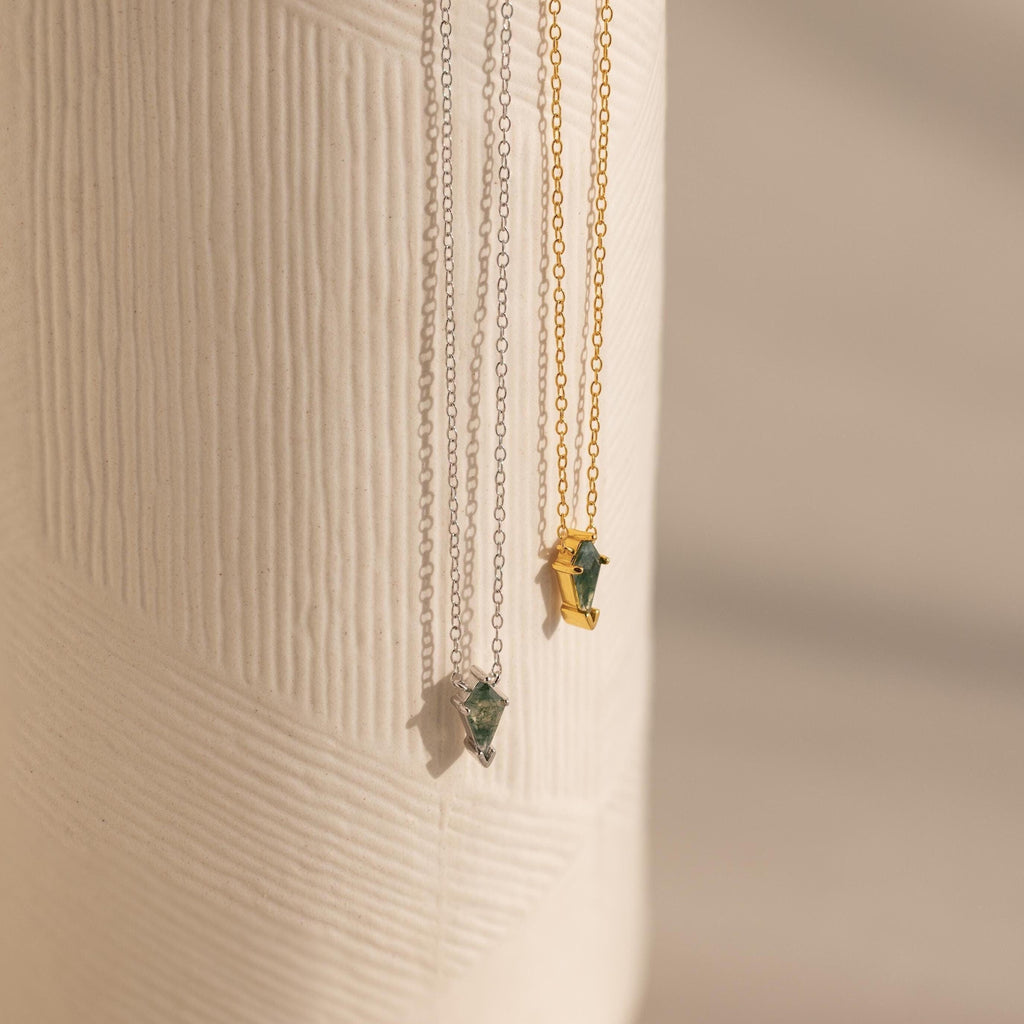 Two Agate Art Deco Pendant Necklaces—one silver, one gold—feature striking green moss agate pendants and hang elegantly against a textured white surface.