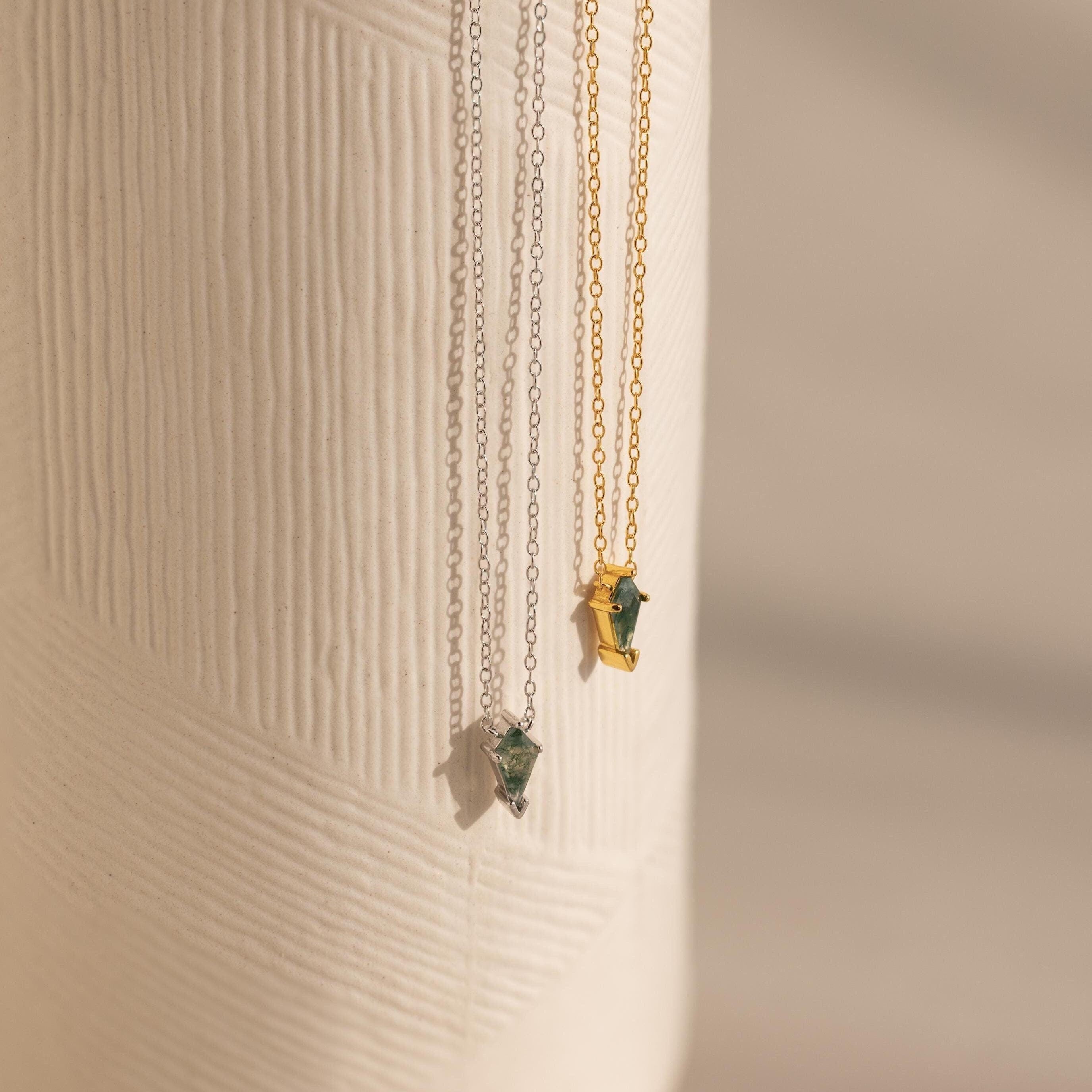 Two Agate Art Deco Pendant Necklaces—one silver, one gold—feature striking green moss agate pendants and hang elegantly against a textured white surface.