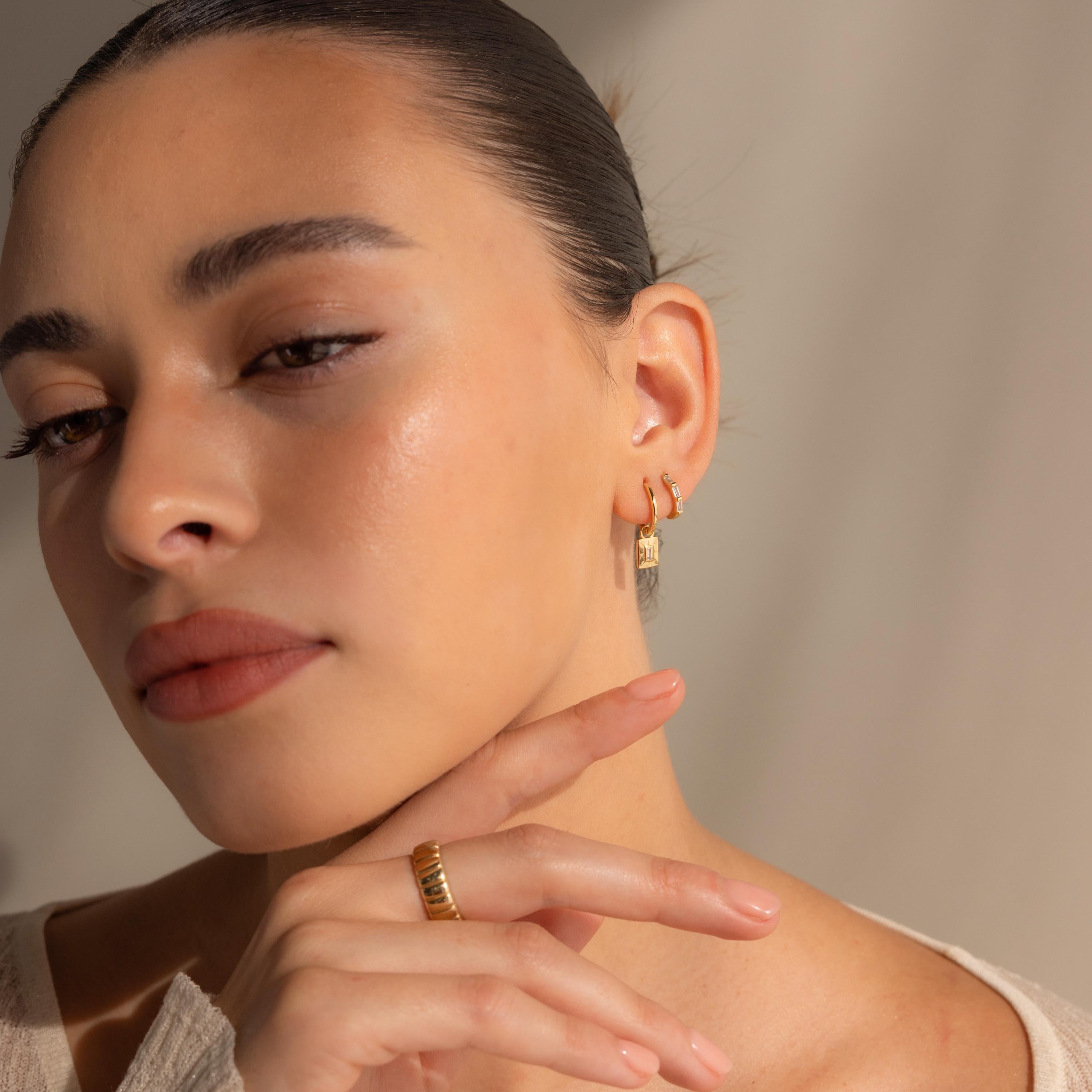 A woman poses for a photo, elegantly showcasing the Art Deco Diamond Tag Huggies—vintage-inspired hoops adorned with sparkling baguette-cut diamonds.