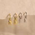 Two pairs of Art Deco Diamond Tag Huggies, one gold and one silver, shown on a textured beige background.