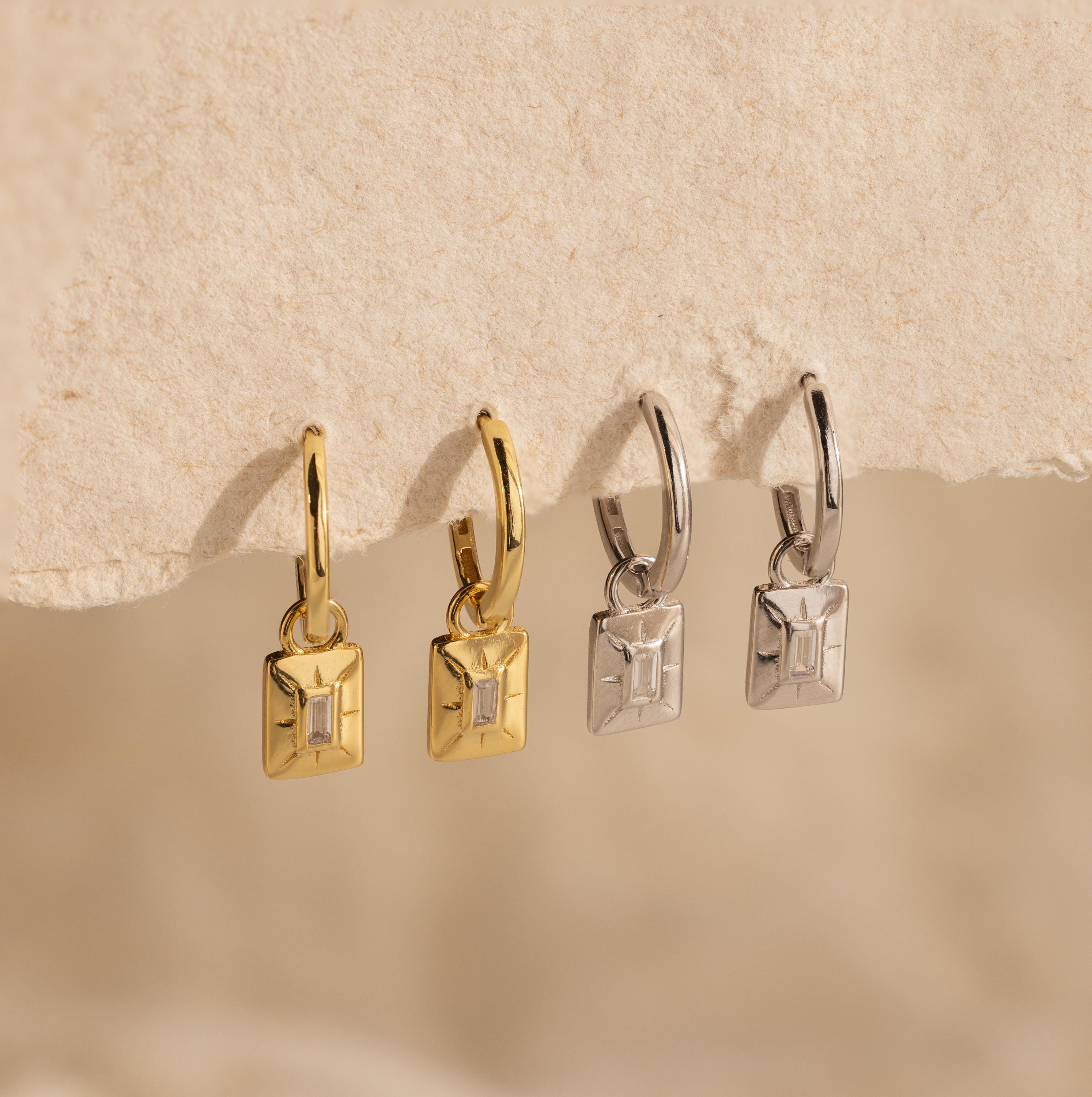 Two pairs of Art Deco Diamond Tag Huggies, one gold and one silver, shown on a textured beige background.