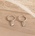 The Art Deco Diamond Tag Huggies, vintage-inspired hoops with geometric pendants, rest on a soft beige surface with lace in the background, reflecting elegant Art Deco design.