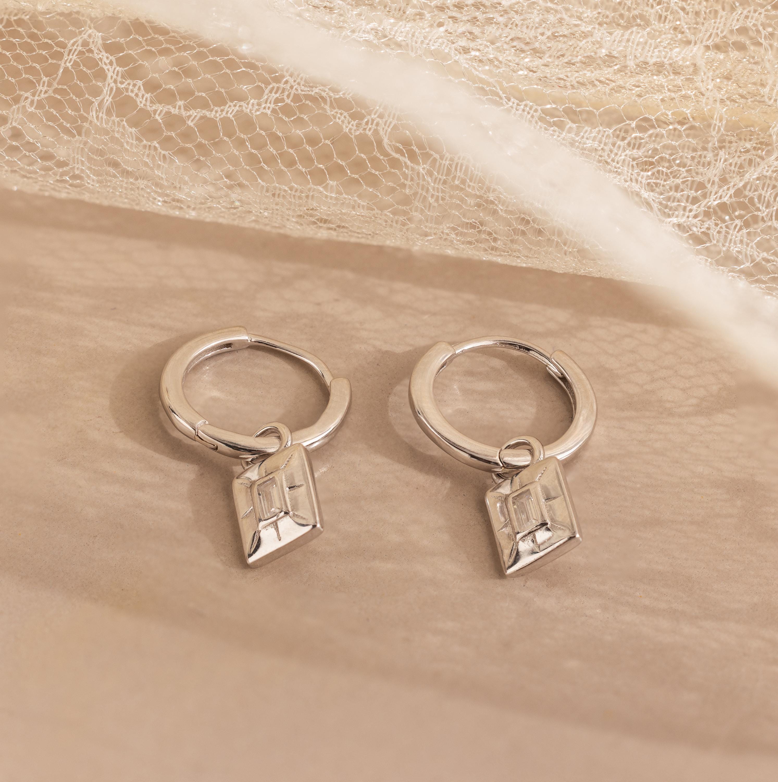 The Art Deco Diamond Tag Huggies, vintage-inspired hoops with geometric pendants, rest on a soft beige surface with lace in the background, reflecting elegant Art Deco design.