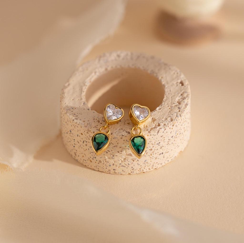 Emerald Heart Drop Studs featuring heart-shaped clear stones and emerald teardrops, displayed on a textured beige stand.