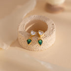 Emerald Heart Drop Studs featuring heart-shaped clear stones and emerald teardrops, displayed on a textured beige stand.