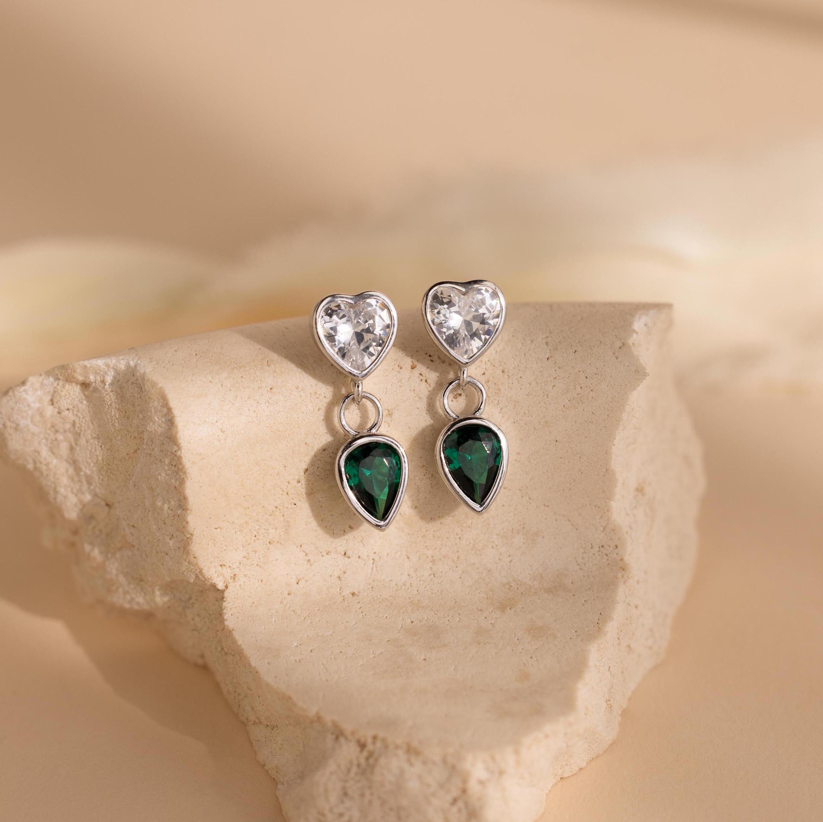Emerald Heart Drop Studs showcase heart-shaped clear stones and vivid green emerald teardrop gems, elegantly presented on a beige stone surface.
