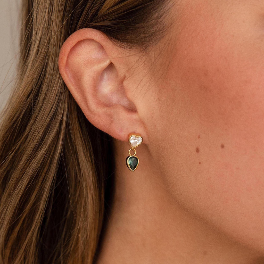 Close-up of a woman's ear wearing Emerald Heart Drop Studs, showcasing a heart-shaped white stone above a green teardrop stone.