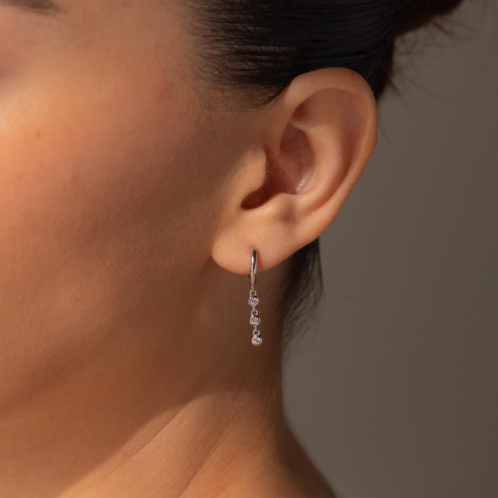 A close-up of a woman's ear adorned with Dangling Diamond Chain Huggies, featuring silver hoops and three sparkling gemstones for an art deco-inspired elegance.