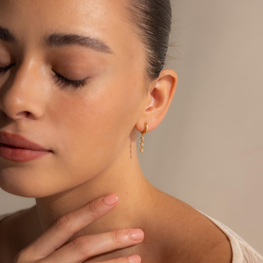 A woman with closed eyes gently touches her neck, wearing Dangling Diamond Chain Huggies that shimmer softly in the light.