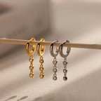 Two pairs of Dangling Diamond Chain Huggies, one gold and one silver, each featuring three dangling crystals in an art deco-inspired design, displayed on a stick.