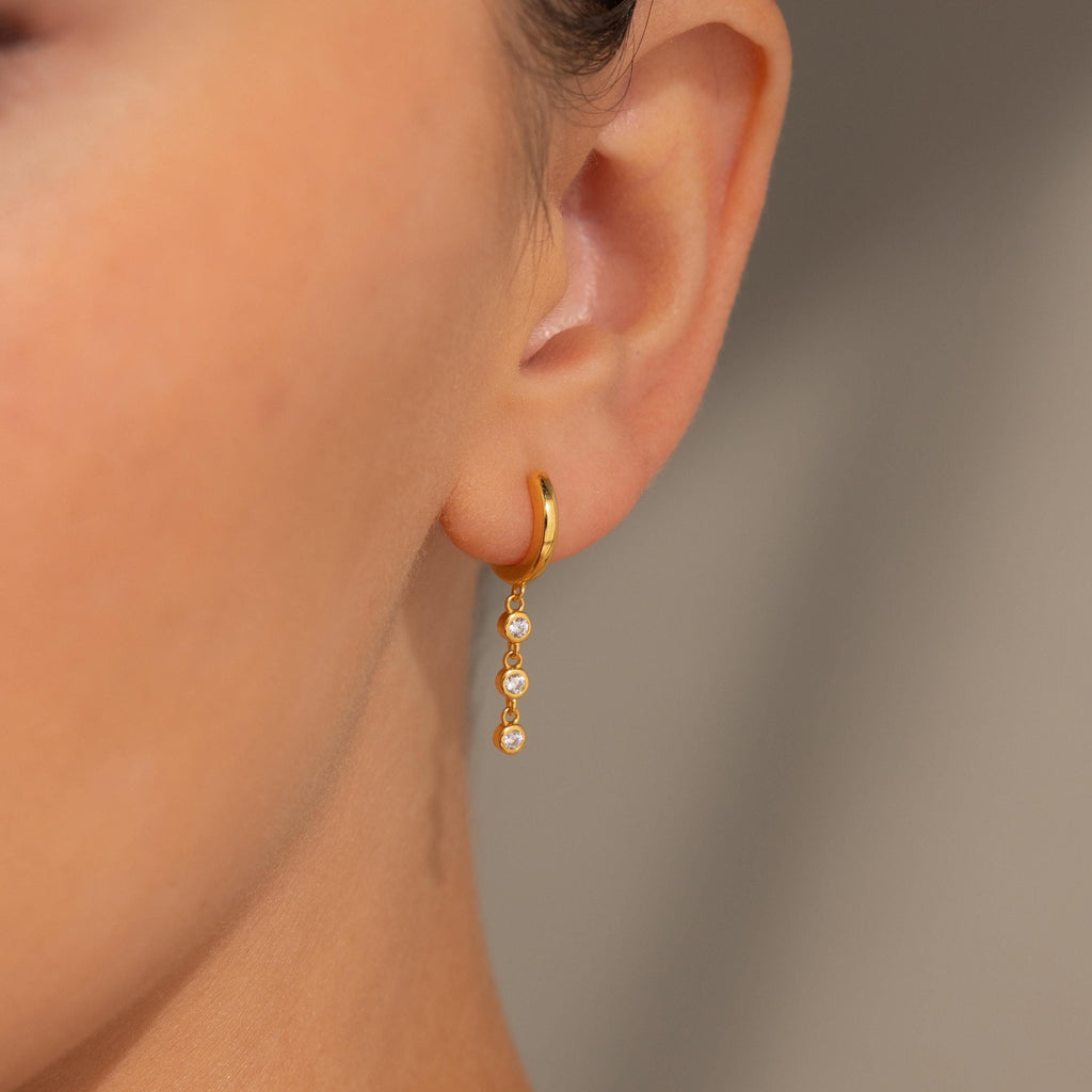 Close-up of an ear wearing Dangling Diamond Chain Huggies, gold hoops featuring three sparkling stones in a design inspired by art deco jewelry.