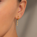 Close-up of an ear wearing Dangling Diamond Chain Huggies, gold hoops featuring three sparkling stones in a design inspired by art deco jewelry.