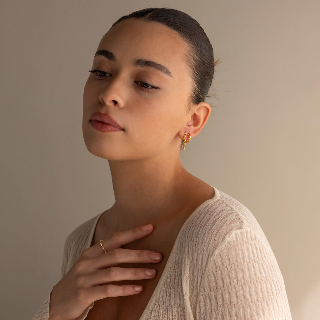 A woman with slicked-back hair wears gold jewelry and Diamond Chain Drop Earrings, paired with a cream ribbed top, gazing thoughtfully downward.