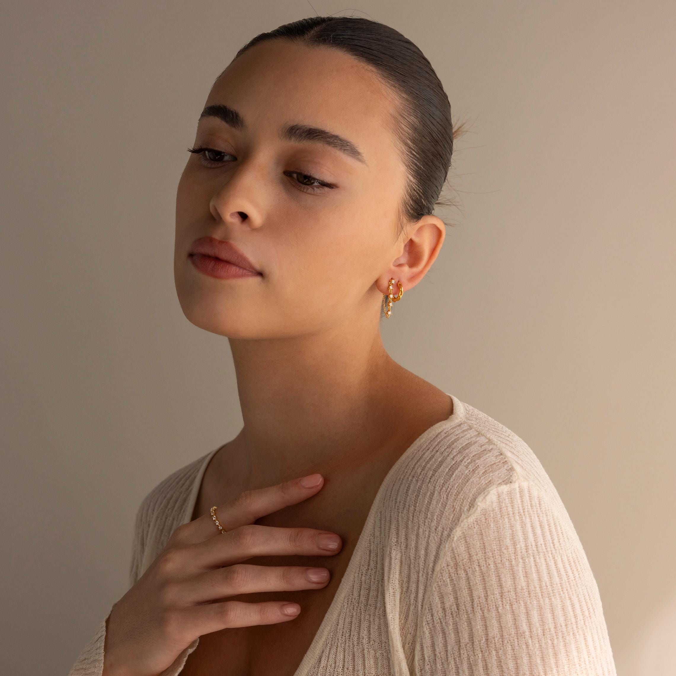 A woman with slicked-back hair wears gold jewelry and Diamond Chain Drop Earrings, paired with a cream ribbed top, gazing thoughtfully downward.