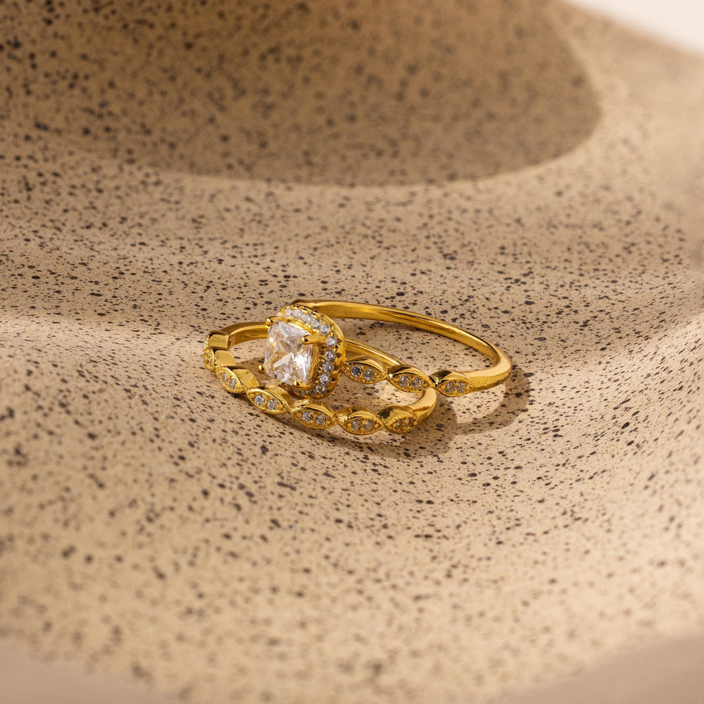 The Vintage Diamond Rings Set features cushion-cut halo stones and marquise-detailed bands, elegantly displayed on a textured beige surface.