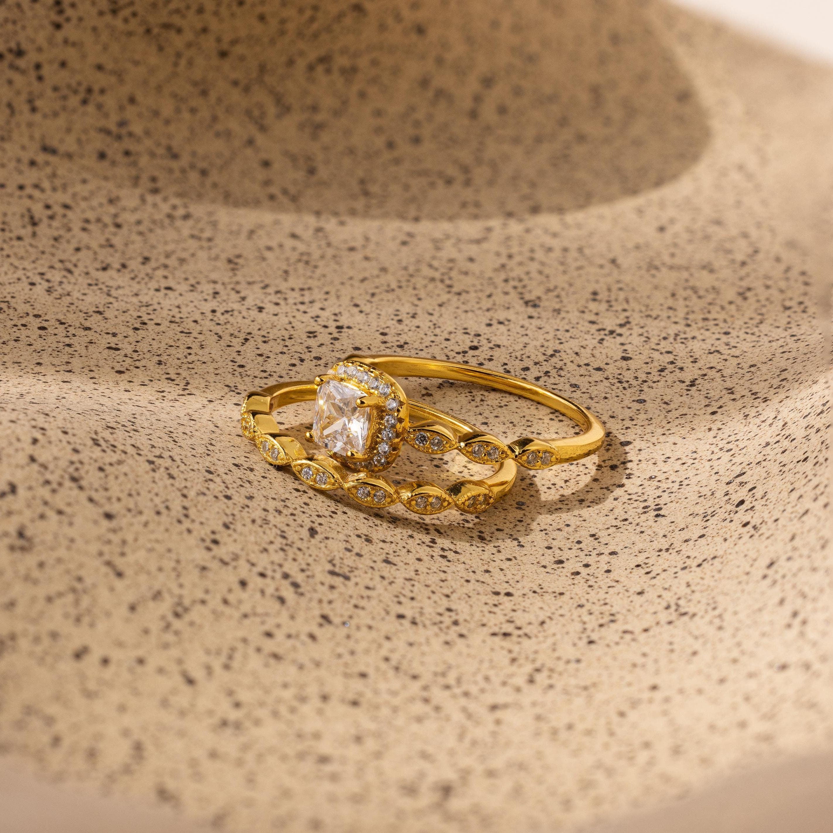 The Vintage Diamond Rings Set features cushion-cut halo stones and marquise-detailed bands, elegantly displayed on a textured beige surface.