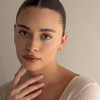 A woman with dark hair in a bun, wearing a cream top and the Agate Kite Drop Earrings, touches her neck while gazing at the camera.