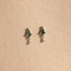 The Agate Kite Drop Earrings feature a geometric silver design with green stones, diamond charms, and dangling crystals, displayed on a beige background.