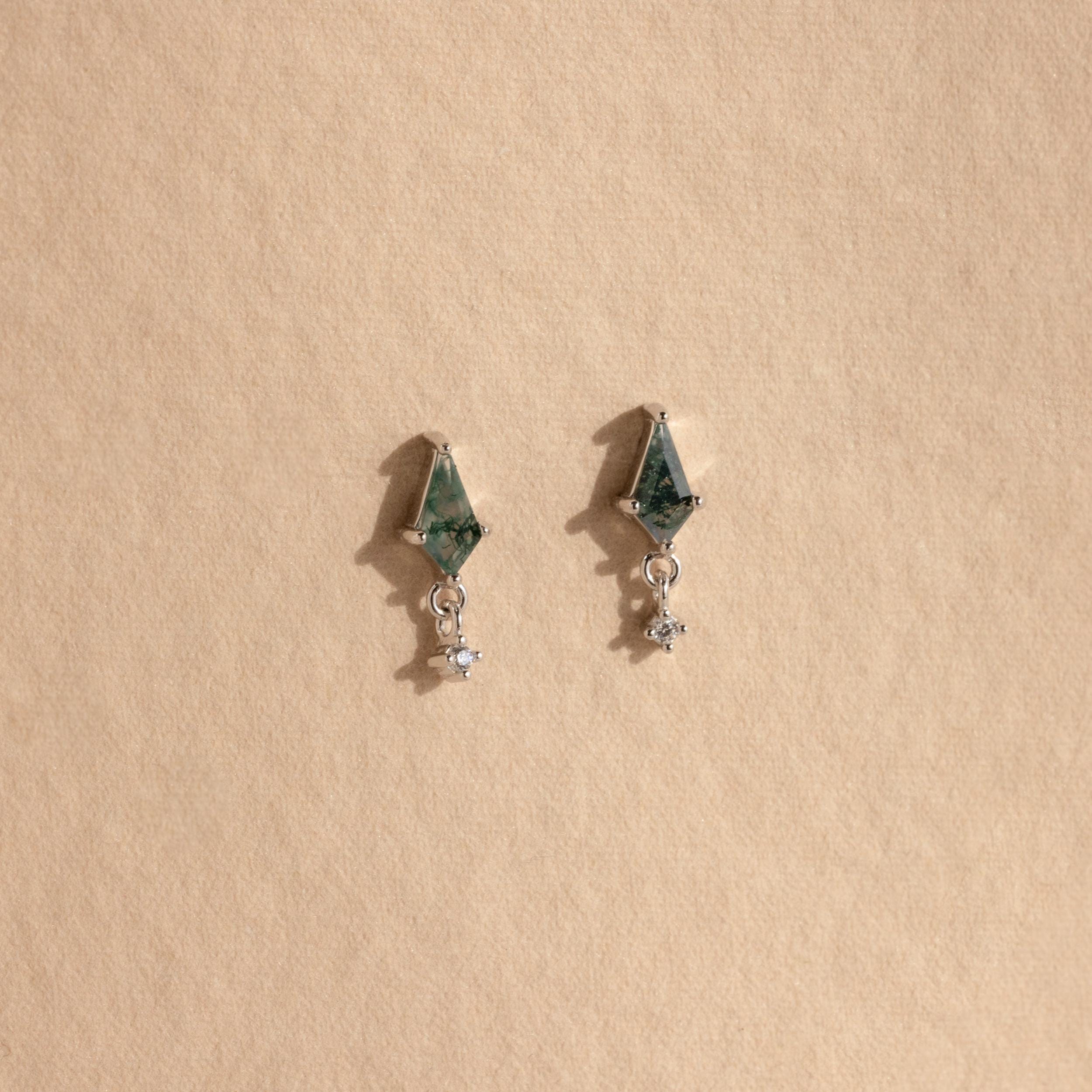 The Agate Kite Drop Earrings feature a geometric silver design with green stones, diamond charms, and dangling crystals, displayed on a beige background.