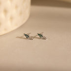 A pair of Agate Kite Drop Earrings with small silver studs and moss agate stones rests on a softly lit beige surface.