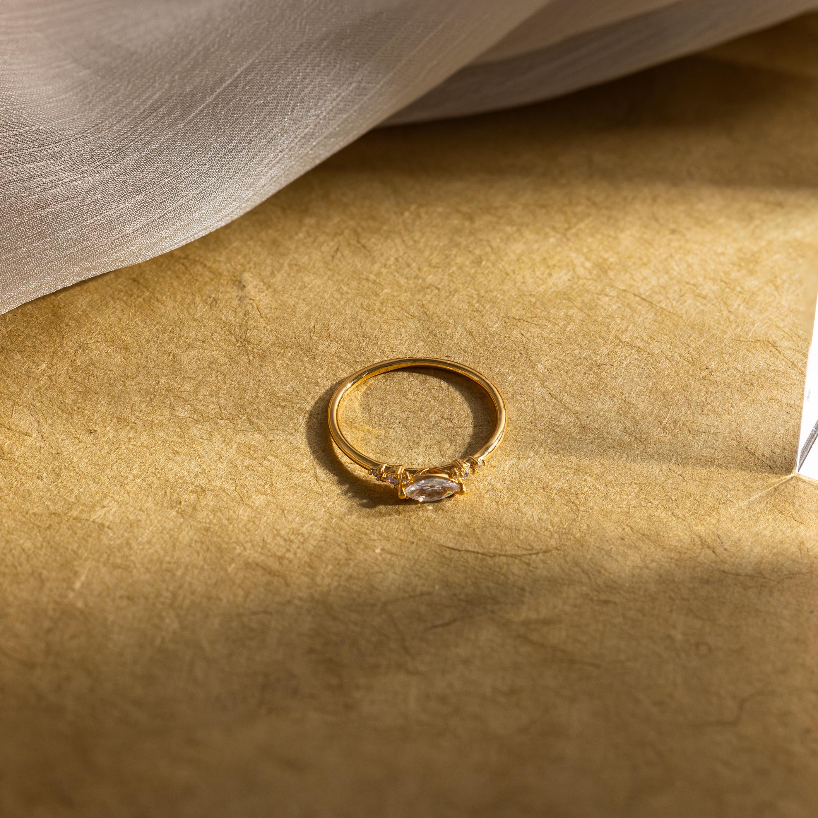 The Marquise Diamond Ring, featuring a gold-finish band, rests on a textured beige surface bathed in gentle sunlight.