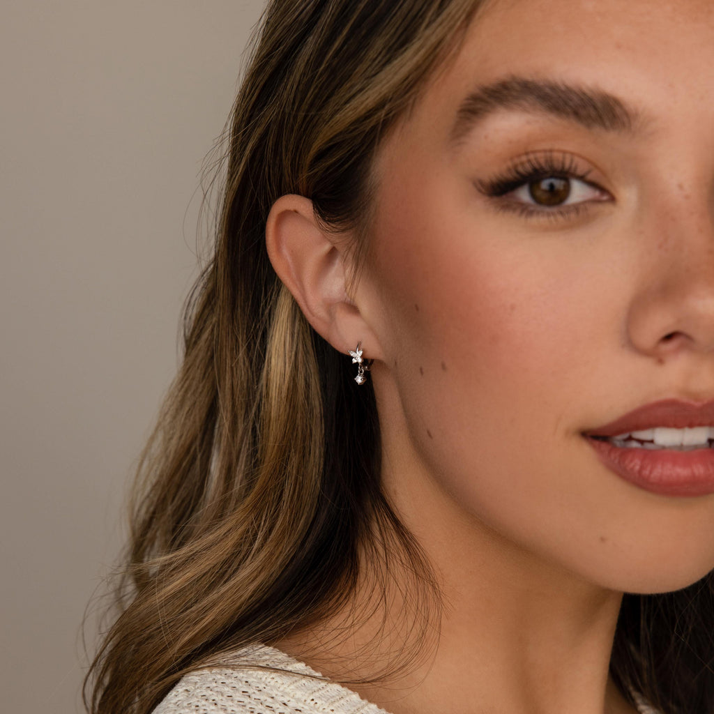 Woman with wavy hair wears elegant Butterfly Drop Huggies in Sterling Silver and a cream knit top, shown in close-up.