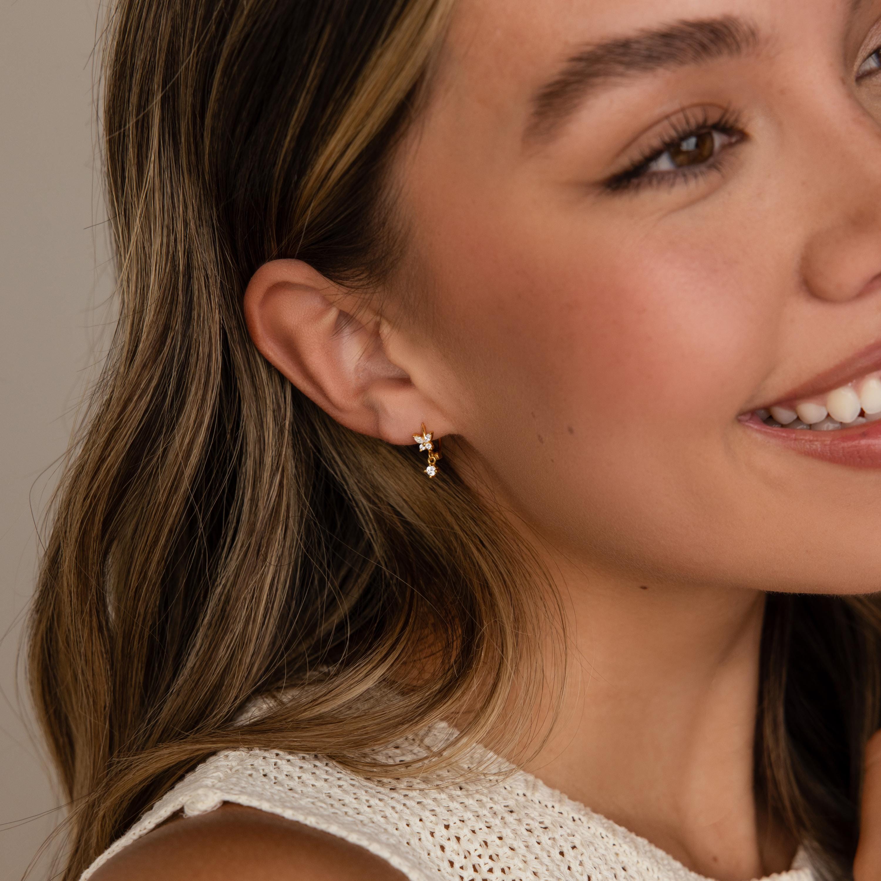 Smiling woman wearing Butterfly Drop Huggies in 18K Gold, featuring delicate butterfly shapes with diamond accents and small dangling gems; close-up on her ear and face.