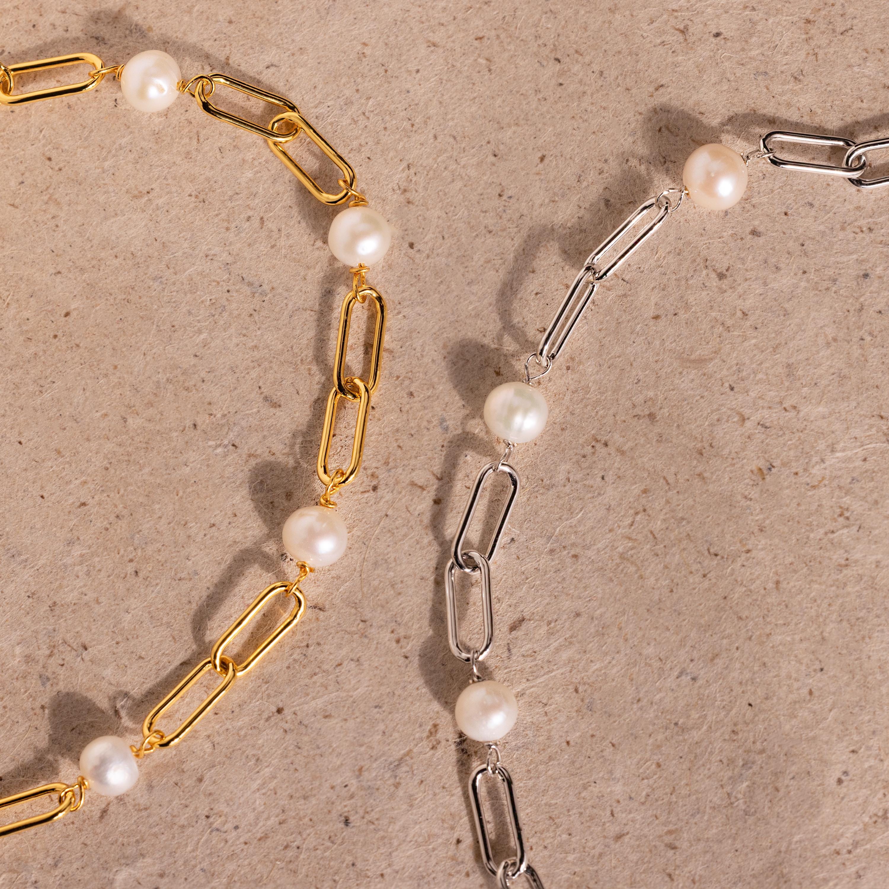 The Pearl Station Paperclip Bracelet is displayed alongside gold and silver chain necklaces with pearl accents on a textured beige surface.
