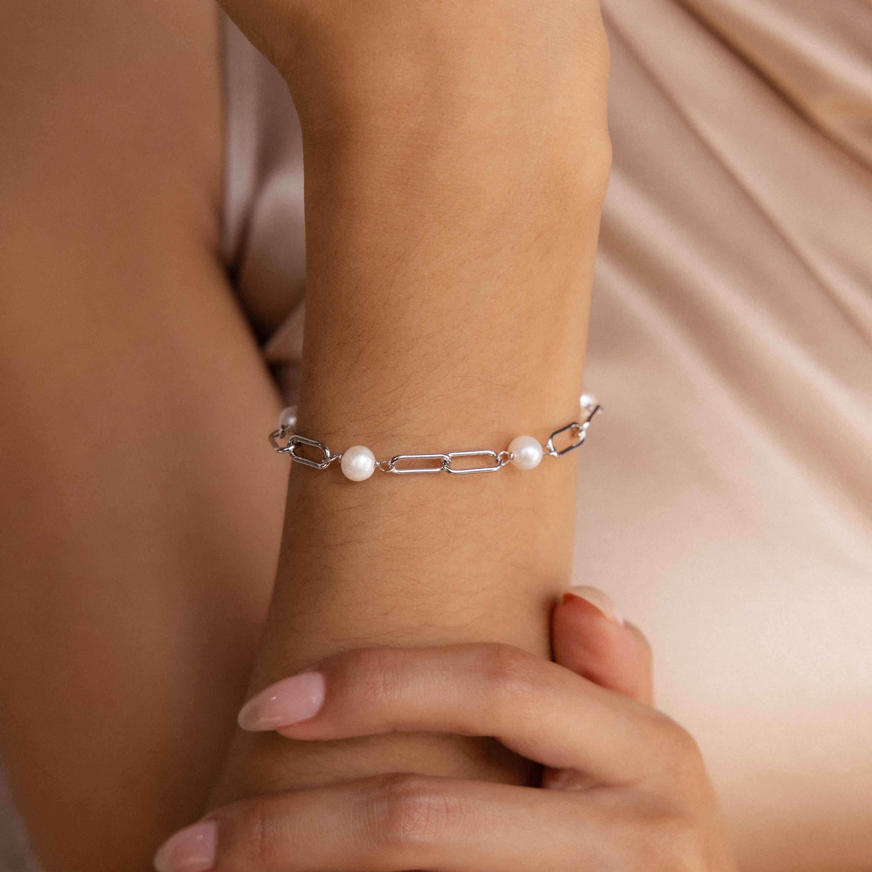 A woman’s wrist showcases the Pearl Station Paperclip Bracelet—silver chain with three pearls—set against a soft beige background.