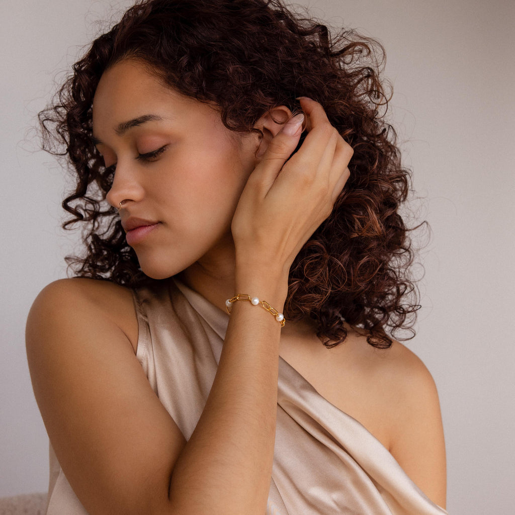 A woman with curly hair, wearing a beige off-shoulder top, gently touches her ear while showcasing the Pearl Station Paperclip Bracelet.