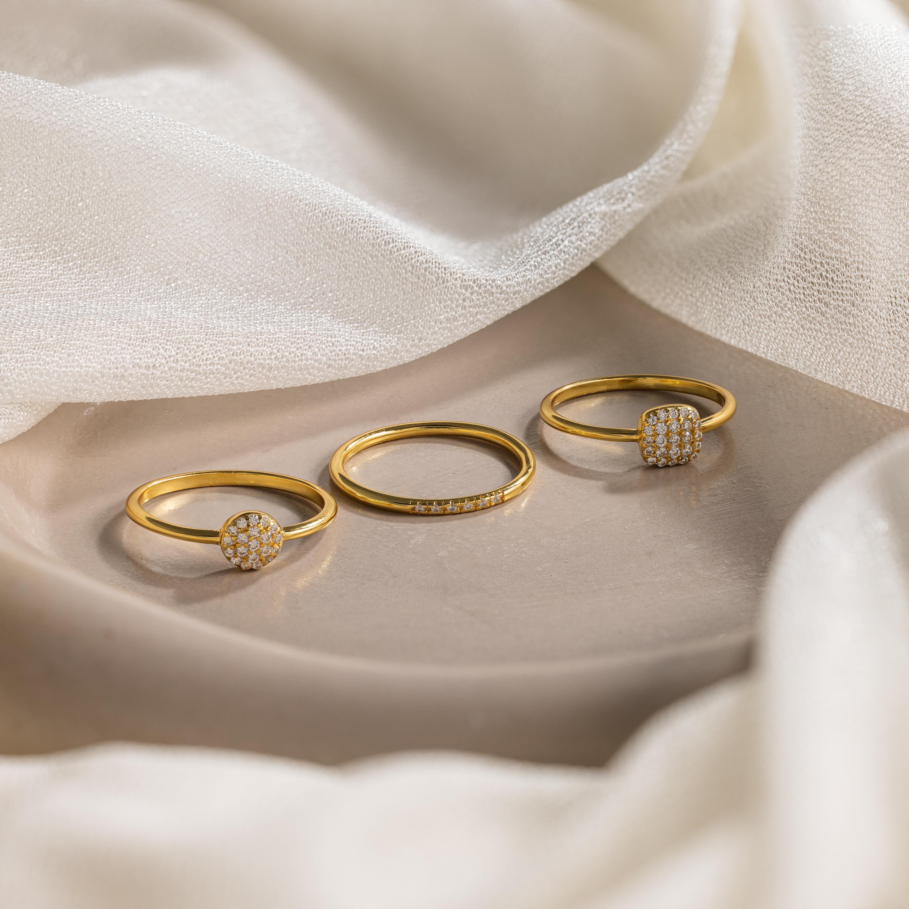The Classic Pave Ring Set, featuring three gold rings with diamond accents, is elegantly displayed on a beige surface with soft white fabric—an ideal jewelry set for any occasion.