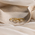 The Classic Pave Ring Set, featuring two gold rings with diamond accents, rests on a speckled beige dish surrounded by soft cream fabric.