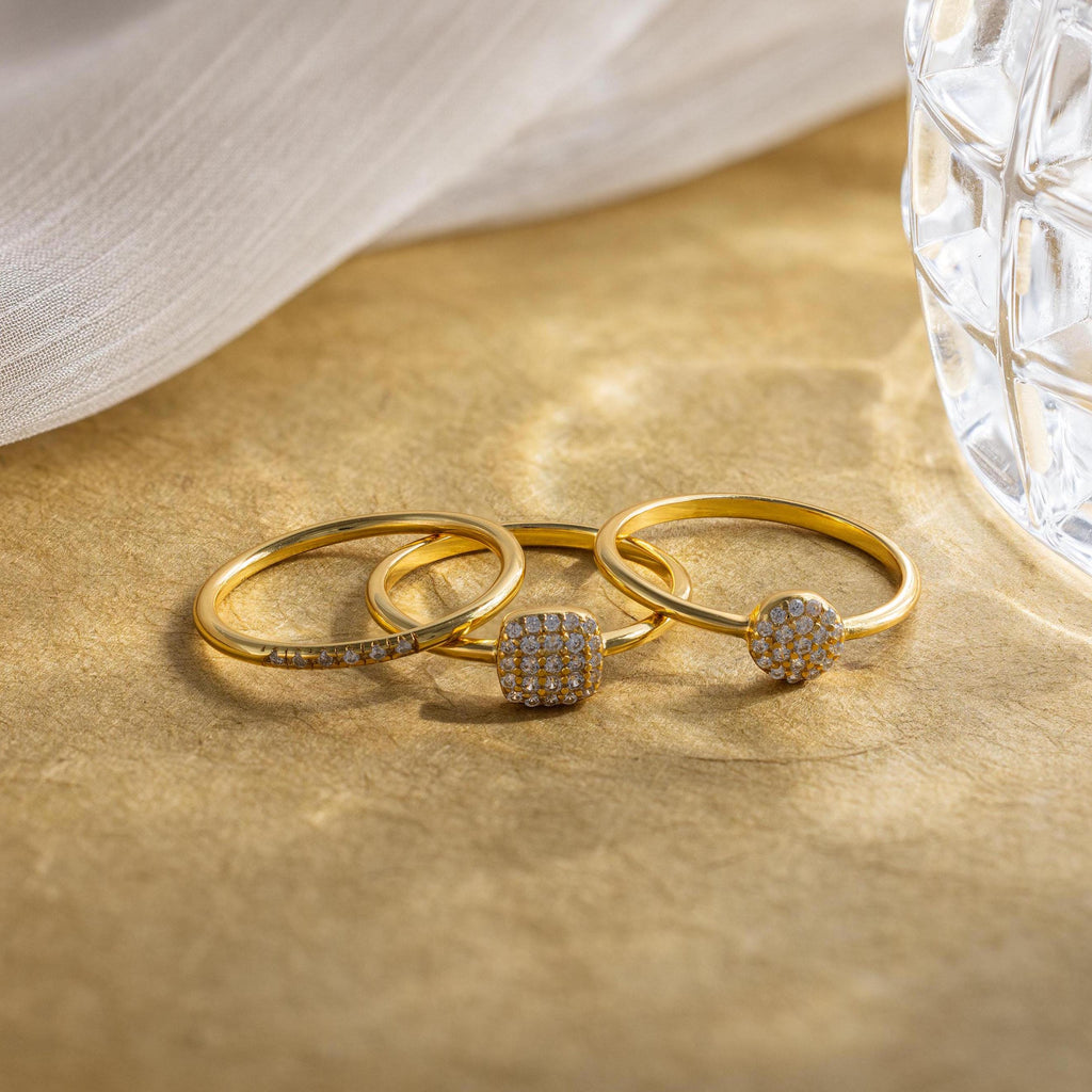 The Classic Pave Ring Set features three elegant gold rings with small diamonds, displayed on a light brown surface near glass and fabric. This luxury set adds sophistication to any look.