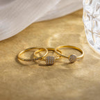 The Classic Pave Ring Set features three elegant gold rings with small diamonds, displayed on a light brown surface near glass and fabric. This luxury set adds sophistication to any look.