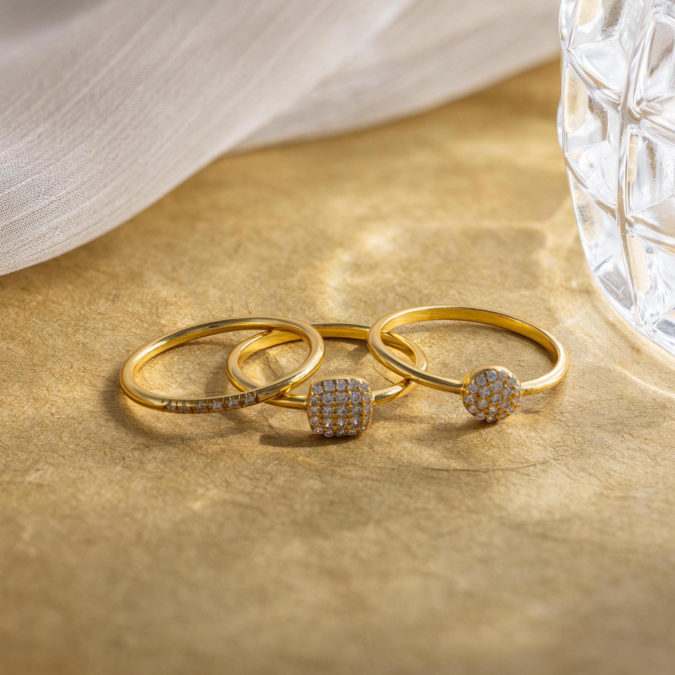 The Classic Pave Ring Set features three elegant gold rings with small diamonds, displayed on a light brown surface near glass and fabric. This luxury set adds sophistication to any look.