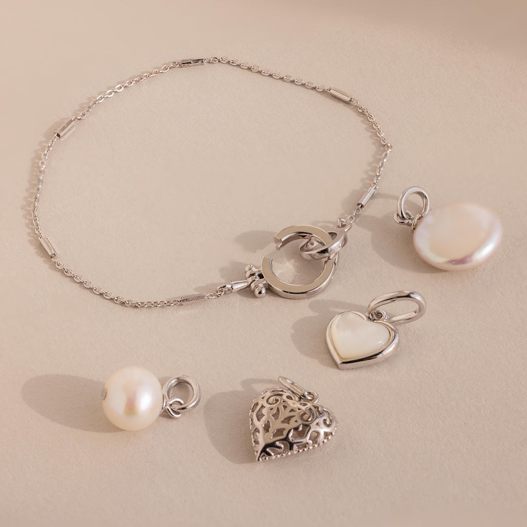 The Hinged Circle Charm Bracelet in Sterling Silver, adorned with pearl and heart charms and paired with two extra pearl and heart pendants, is beautifully displayed on a beige surface.