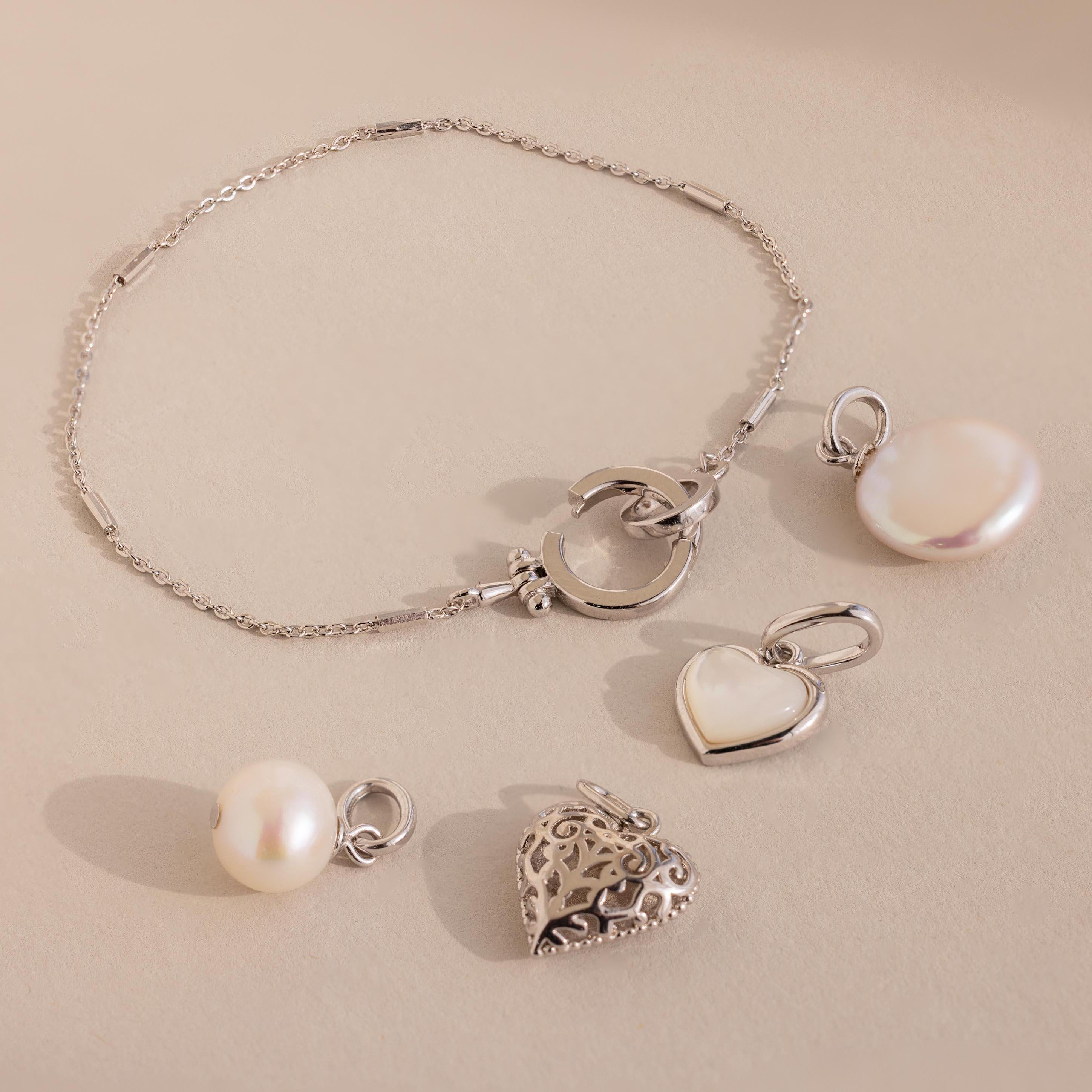 The Hinged Circle Charm Bracelet in Sterling Silver, adorned with pearl and heart charms and paired with two extra pearl and heart pendants, is beautifully displayed on a beige surface.
