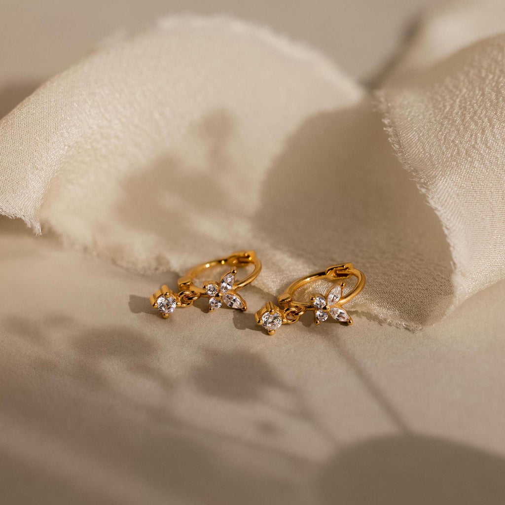 Butterfly Drop Huggies in 18K Gold, featuring delicate butterfly charms and sparkling accents, displayed on a soft cream fabric background.