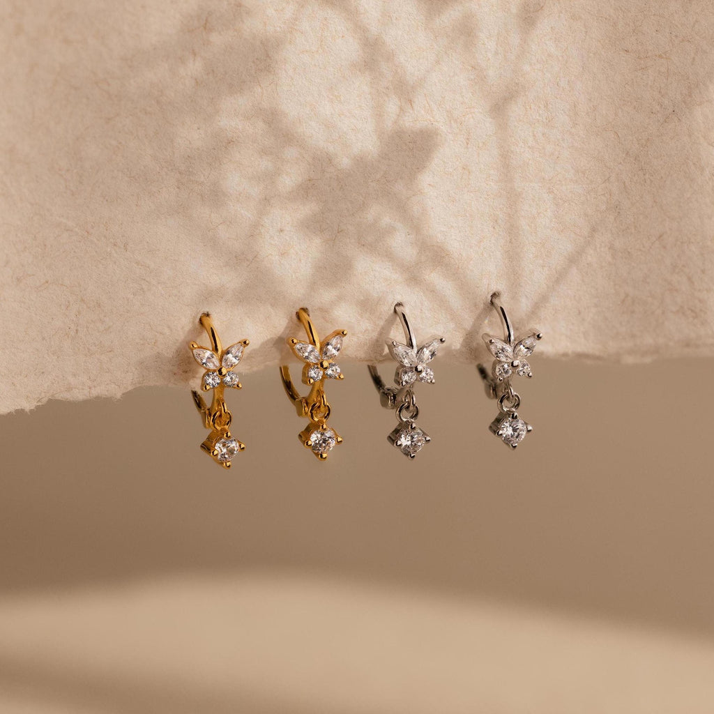 Two pairs of small, flower-shaped earrings, including delicate Butterfly Drop Huggies in gold and silver, are displayed on textured beige paper.