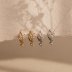 Two pairs of small, flower-shaped earrings, including delicate Butterfly Drop Huggies in gold and silver, are displayed on textured beige paper.
