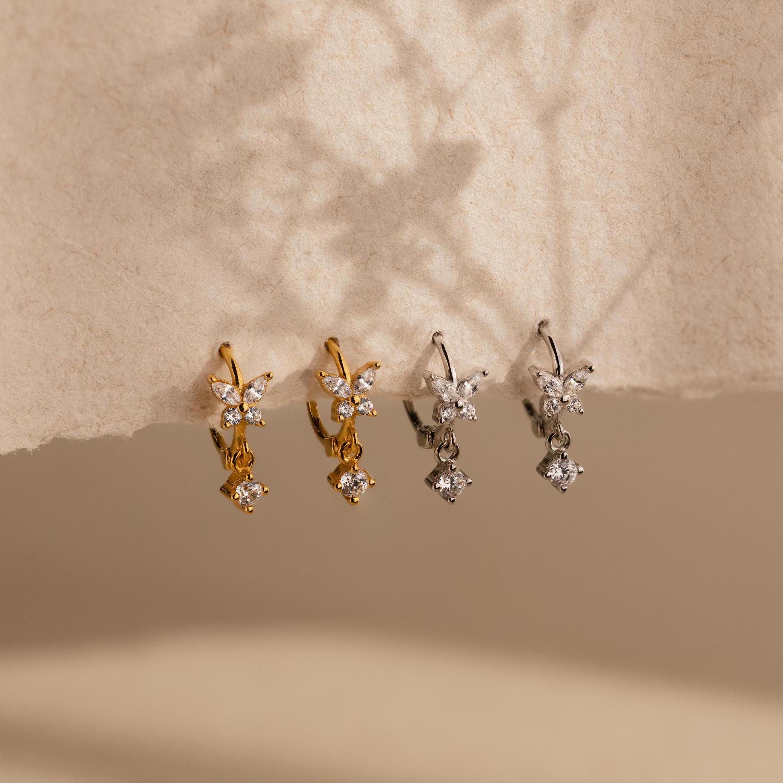 Two pairs of small, flower-shaped earrings, including delicate Butterfly Drop Huggies in gold and silver, are displayed on textured beige paper.